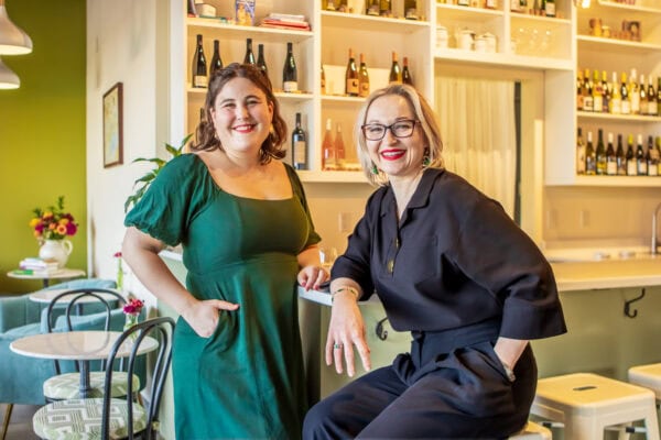 Two women smiling at a wine bar with shelves of wine bottles behind them.